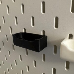 以下が含まれることがあります： 黒色の長方形の収納ビン。ペグボードに取り付けるように設計されています。白いペグボードに取り付けられており、楕円形の穴が等間隔に配置されています。白いビンも表示されています。