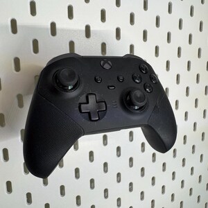 May include: A black Xbox controller with textured grips and various buttons, including a directional pad and analogue sticks. The controller is mounted on a white pegboard with evenly spaced holes.