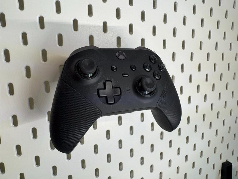 May include: A black Xbox controller with textured grips and various buttons, including a directional pad and analogue sticks. The controller is mounted on a white pegboard with evenly spaced holes.