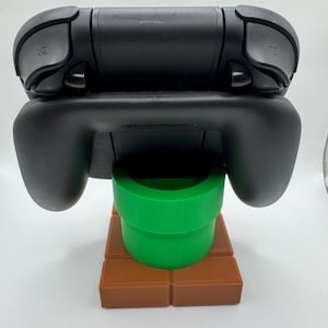 May include: Black Nintendo Switch controller on a green pipe and brown brick base. The controller has ZR and ZL buttons. The base is a stylized representation of a video game level.