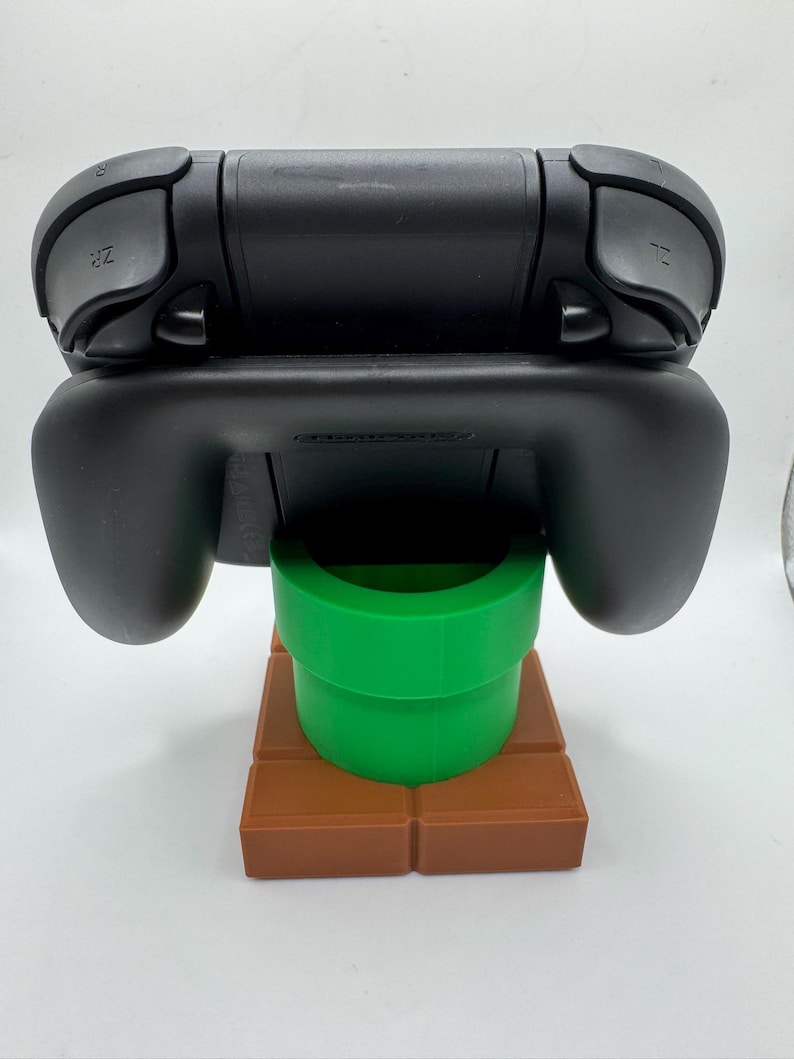 May include: Black Nintendo Switch controller on a green pipe and brown brick base. The controller has ZR and ZL buttons. The base is a stylized representation of a video game level.