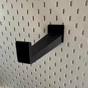 K&ouml;nnte beinhalten: Ein schwarzer, abgewinkelter Regalhalter f&uuml;r ein Lochwandsystem. Der Halter besteht aus einem soliden, mattschwarzen Material und verf&uuml;gt &uuml;ber eine rechteckige Basis und ein leicht abgewinkeltes Regal. Die Lochwand ist wei&szlig; mit gleichm&auml;&szlig;ig verteilten ovalen L&ouml;chern.