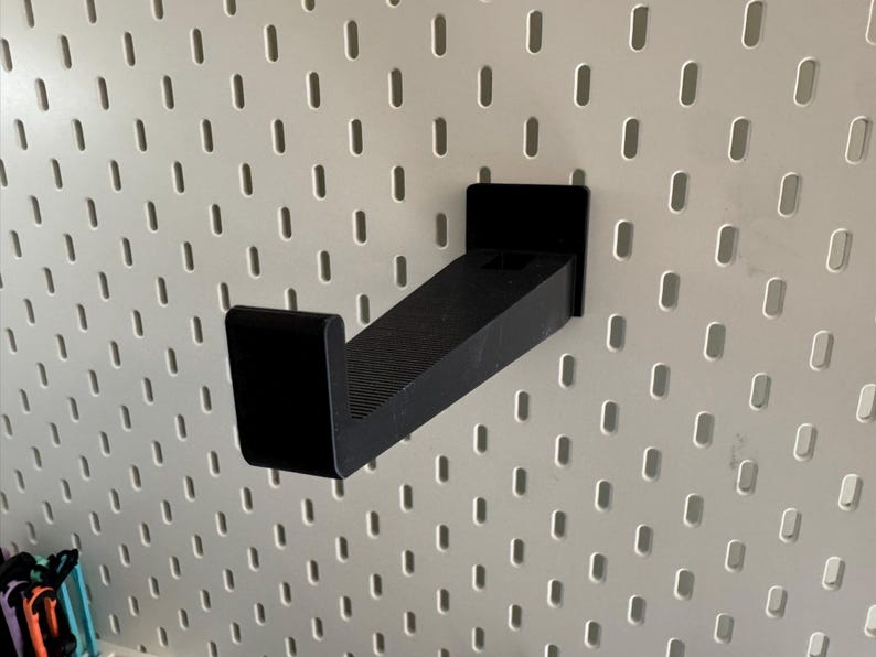 K&ouml;nnte beinhalten: Ein schwarzer, abgewinkelter Regalhalter f&uuml;r ein Lochwandsystem. Der Halter besteht aus einem soliden, mattschwarzen Material und verf&uuml;gt &uuml;ber eine rechteckige Basis und ein leicht abgewinkeltes Regal. Die Lochwand ist wei&szlig; mit gleichm&auml;&szlig;ig verteilten ovalen L&ouml;chern.