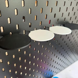 Op de afbeelding: Drie ronde planken, twee wit en een zwart, zijn gemonteerd op een zwarte prikbordwand. De planken zijn gemaakt van glad, hard plastic.