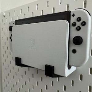 Könnte beinhalten: Eine weiße Nintendo Switch-Konsole und Controller sind mit schwarzen Halterungen an einer weißen Lochwand befestigt. Die Switch befindet sich in einer Dockingstation. Die Controller haben schwarze Knöpfe und Joysticks.