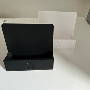 May include: A black rectangular box with a hinged lid and a small cutout on the front, next to a white rectangular stand. Both items are on a white surface. The black box has a small, stylized design on the front.