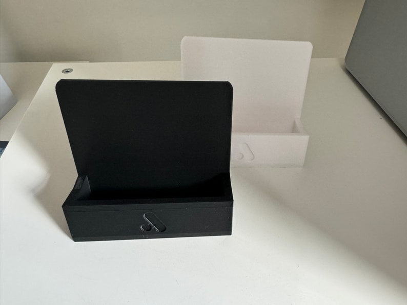 May include: A black rectangular box with a hinged lid and a small cutout on the front, next to a white rectangular stand. Both items are on a white surface. The black box has a small, stylized design on the front.