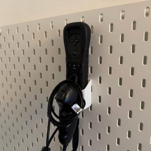 May include: Black Wii remote and nunchuck controller set in a white holder, mounted on a white pegboard. The remote has a black silicone sleeve. The pegboard has evenly spaced oval holes.