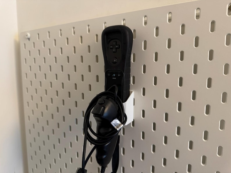 May include: Black Wii remote and nunchuck controller set in a white holder, mounted on a white pegboard. The remote has a black silicone sleeve. The pegboard has evenly spaced oval holes.