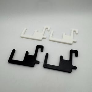 May include: Four plastic brackets, two in white and two in black, are displayed against a white backdrop. Each bracket features a rectangular base with a hook extending from one end and a shorter hook from the other.