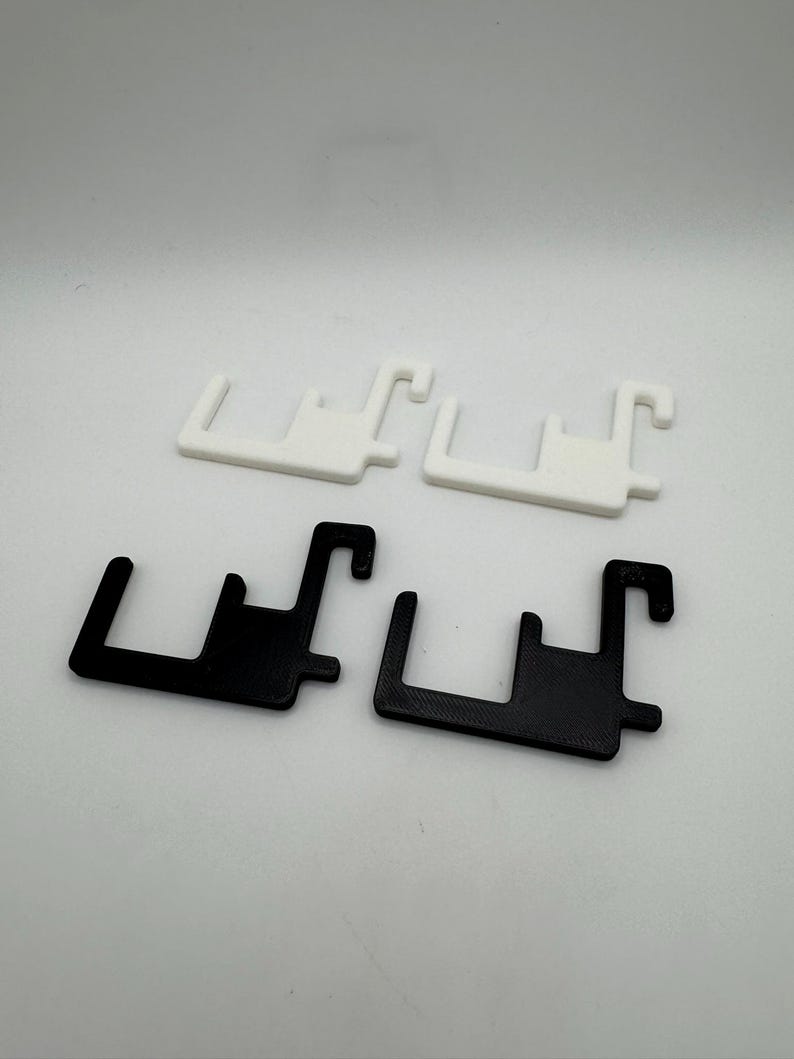 May include: Four plastic brackets, two in white and two in black, are displayed against a white backdrop. Each bracket features a rectangular base with a hook extending from one end and a shorter hook from the other.