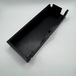 May include: A black plastic tray with a rectangular shape and a raised lip around the edge. The tray has a series of small, rectangular cutouts along the sides.