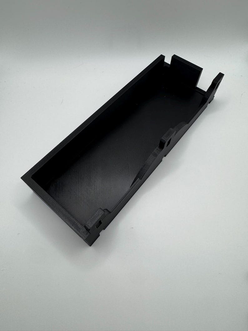 May include: A black plastic tray with a rectangular shape and a raised lip around the edge. The tray has a series of small, rectangular cutouts along the sides.