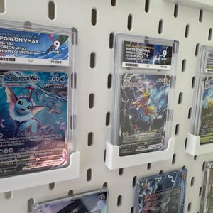 Może przedstawiać: Dziewięć kart kolekcjonerskich Pokemon jest wyświetlanych w ochronnych etui na białej ścianie z tablicą perforowaną. Karty przedstawiają Vaporeon VMAX, Jolteon VMAX i Flareon VMAX, wszystkie z serii Black Star Promo z 2021 roku. Karty są oceniane na 9 z 10 za stan.