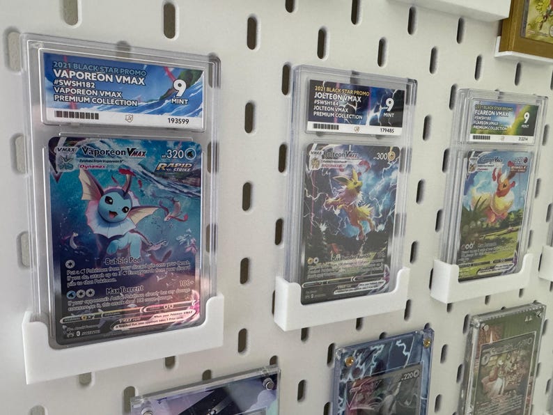 Może przedstawiać: Dziewięć kart kolekcjonerskich Pokemon jest wyświetlanych w ochronnych etui na białej ścianie z tablicą perforowaną. Karty przedstawiają Vaporeon VMAX, Jolteon VMAX i Flareon VMAX, wszystkie z serii Black Star Promo z 2021 roku. Karty są oceniane na 9 z 10 za stan.