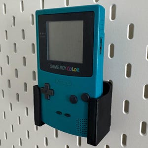 May include: A teal Game Boy Color handheld video game console with a black screen and a black plastic wall mount. The console is mounted on a white pegboard.