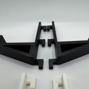 May include: Black 3D-printed stand with a triangular base and a rectangular support structure. The stand is designed to hold an item. Two white 3D-printed support pieces are also visible.
