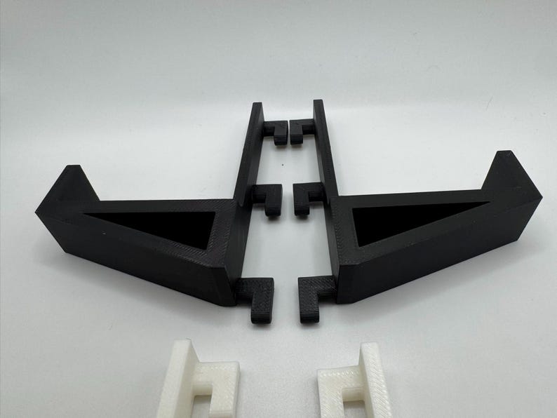 May include: Black 3D-printed stand with a triangular base and a rectangular support structure. The stand is designed to hold an item. Two white 3D-printed support pieces are also visible.