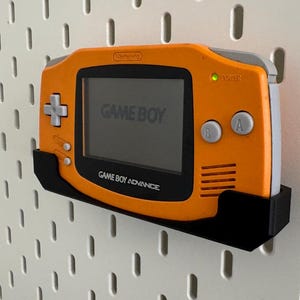 Op de afbeelding: Een oranje Game Boy Advance handheld-videogamesysteem met een zwarte wandbevestiging. De console heeft een grijs scherm met de tekst "GAME BOY" erop. De console is gemonteerd op een witte prikbordwand.