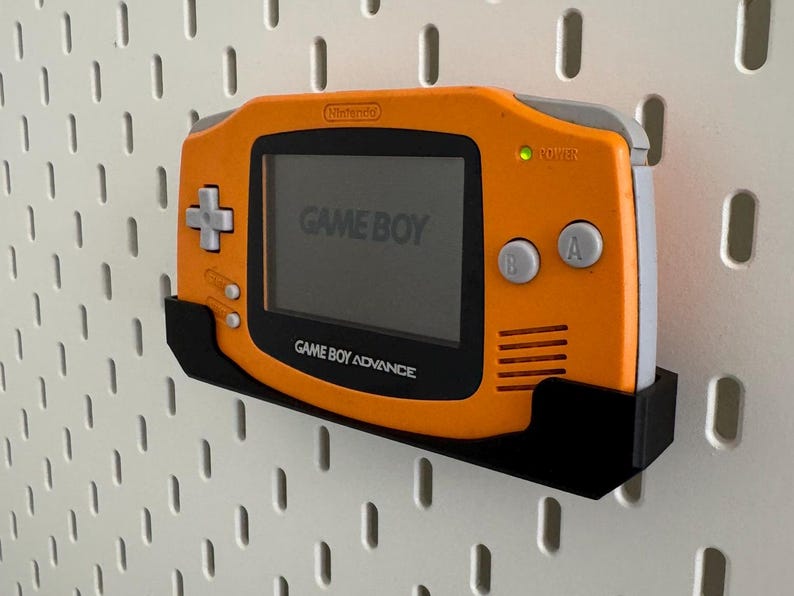 Op de afbeelding: Een oranje Game Boy Advance handheld-videogamesysteem met een zwarte wandbevestiging. De console heeft een grijs scherm met de tekst "GAME BOY" erop. De console is gemonteerd op een witte prikbordwand.
