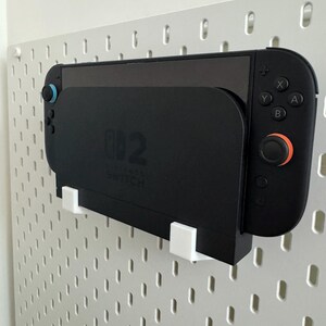 Op de afbeelding: Een zwarte Nintendo Switch-console en controllers zijn gemonteerd op een wit pegboard. De console zit in een zwart oplaadstation. De controllers hebben blauwe en oranje accenten. Het pegboard heeft een patroon van gaten.