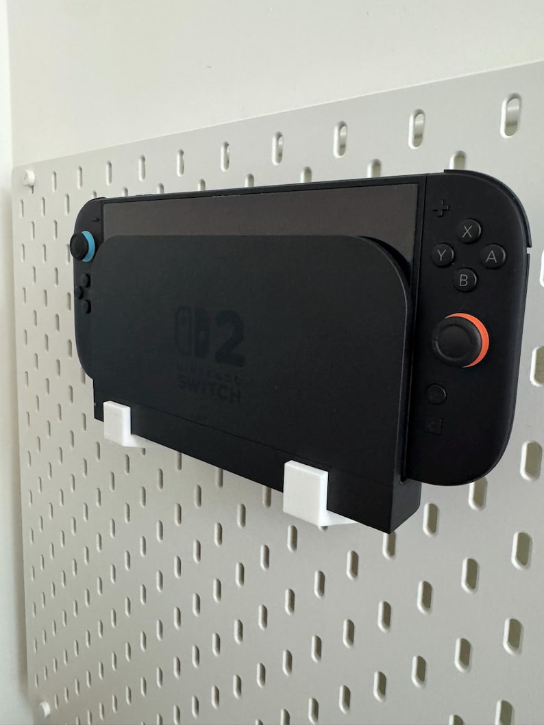 Op de afbeelding: Een zwarte Nintendo Switch-console en controllers zijn gemonteerd op een wit pegboard. De console zit in een zwart oplaadstation. De controllers hebben blauwe en oranje accenten. Het pegboard heeft een patroon van gaten.