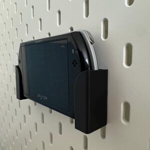 Pu&ograve; includere: Console di gioco portatile PSP nera in un supporto a parete nero, fissata a una parete forata bianca. La console &egrave; etichettata "SONY" e "PSP". Il supporto &egrave; progettato per tenere saldamente la console.