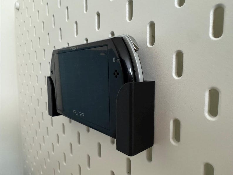 Pu&ograve; includere: Console di gioco portatile PSP nera in un supporto a parete nero, fissata a una parete forata bianca. La console &egrave; etichettata "SONY" e "PSP". Il supporto &egrave; progettato per tenere saldamente la console.