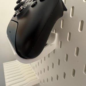 May include: A black video game controller mounted on a white pegboard using a white 3D-printed holder. The controller is angled, showing the buttons and joysticks. The pegboard has evenly spaced, oblong holes.