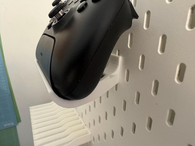May include: A black video game controller mounted on a white pegboard using a white 3D-printed holder. The controller is angled, showing the buttons and joysticks. The pegboard has evenly spaced, oblong holes.