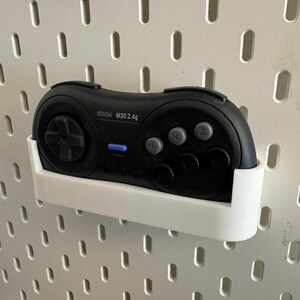 Könnte beinhalten: Ein schwarzes M30 2.4g Wireless-Gamepad liegt in einer weißen Halterung, die an einer weißen Lochwand befestigt ist. Das Gamepad hat ein Steuerkreuz, graue Tasten und eine blaue Betriebsanzeige. Die Lochwand hat gleichmäßig verteilte ovale Löcher.