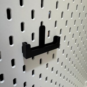 May include: A black pegboard hook is mounted on a white pegboard. The hook is designed to hold items and is made of a sturdy material. The pegboard has evenly spaced oval holes.