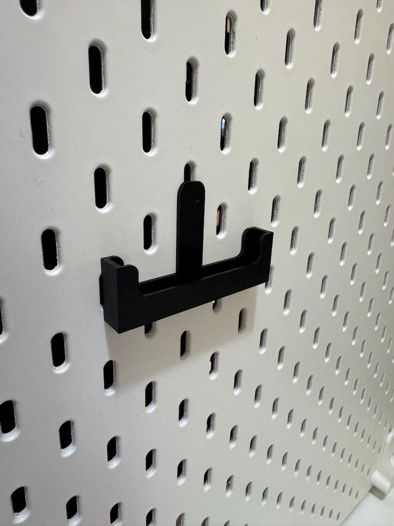 May include: A black pegboard hook is mounted on a white pegboard. The hook is designed to hold items and is made of a sturdy material. The pegboard has evenly spaced oval holes.