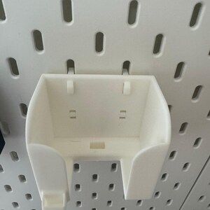May include: A white plastic pegboard accessory with a curved front and two side supports. The accessory is designed to attach to a pegboard with a series of small holes. The accessory is designed to hold small items.