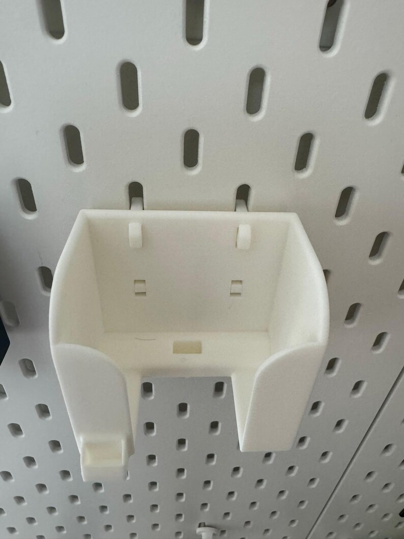 May include: A white plastic pegboard accessory with a curved front and two side supports. The accessory is designed to attach to a pegboard with a series of small holes. The accessory is designed to hold small items.