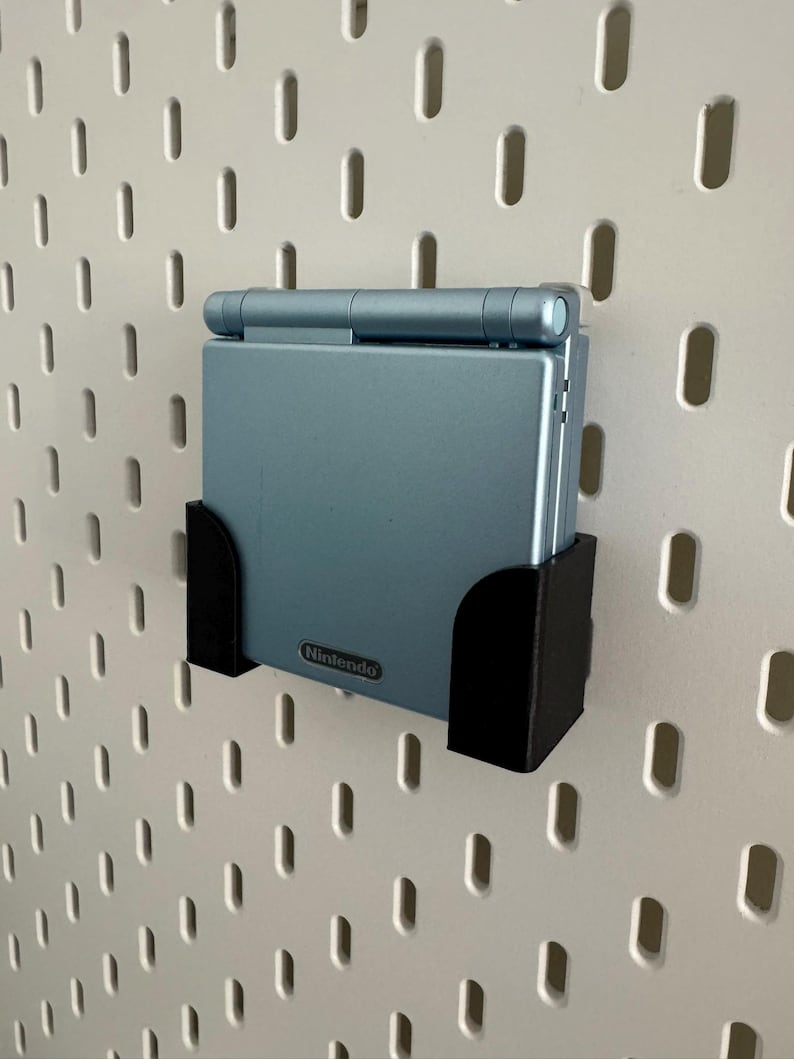 Pu&ograve; includere: Un supporto da parete in plastica nera per una console di gioco portatile Nintendo Game Boy Advance SP blu chiaro. Il supporto &egrave; fissato a una bacheca bianca.