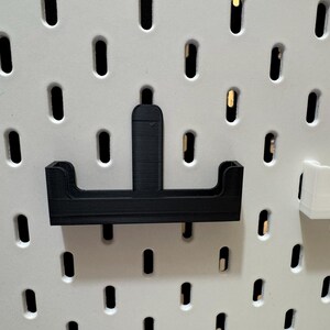 May include: A black 3D-printed pegboard accessory, designed to hold items. The accessory has a horizontal base with two raised sides and a vertical support. It is mounted on a white pegboard with evenly spaced holes.