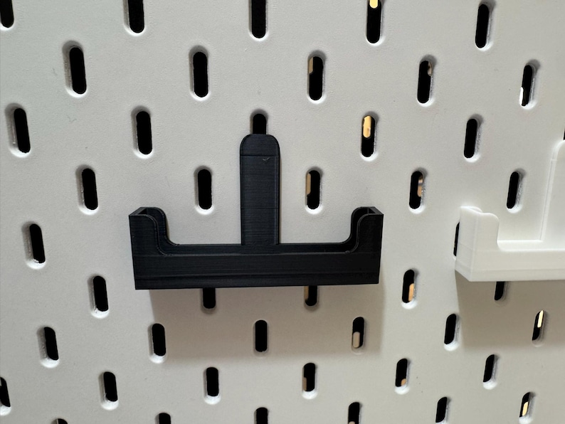 May include: A black 3D-printed pegboard accessory, designed to hold items. The accessory has a horizontal base with two raised sides and a vertical support. It is mounted on a white pegboard with evenly spaced holes.