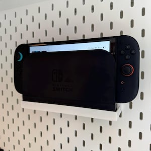 May include: A black Nintendo Switch console docked in a white wall-mounted holder. The screen displays a user interface. The holder is mounted on a white pegboard wall. The Switch has a black body with blue and orange accents.