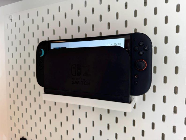 Puede incluir: Una consola Nintendo Switch negra acoplada en un soporte blanco montado en la pared. La pantalla muestra una interfaz de usuario. El soporte est&aacute; montado en una pared de tablero perforado blanco. La Switch tiene un cuerpo negro con detalles en azul y naranja.