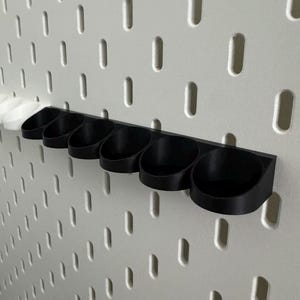 以下が含まれることがあります： 黒色の3Dプリントオーガナイザー。6つのカップ型ホルダーが水平バーに取り付けられています。オーガナイザーは、等間隔の穴が開いた白いペグボードに取り付けられています。ホルダーは、小さなアイテムを収納するように設計されています。