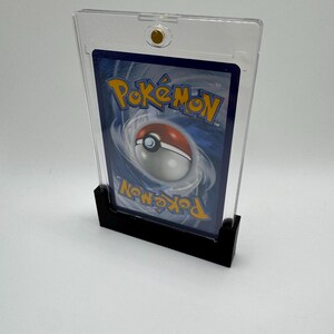 Puede incluir: Un soporte de tarjeta de pl&aacute;stico transparente con una base negra, que muestra el reverso de una tarjeta coleccionable de Pok&eacute;mon. La tarjeta presenta un fondo azul con el logotipo de Pok&eacute;mon y un dise&ntilde;o de Pok&eacute; Ball. El soporte tiene un cierre dorado.