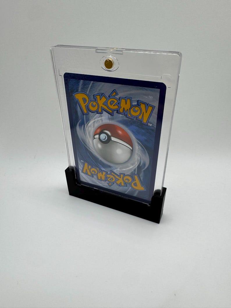 Puede incluir: Un soporte de tarjeta de pl&aacute;stico transparente con una base negra, que muestra el reverso de una tarjeta coleccionable de Pok&eacute;mon. La tarjeta presenta un fondo azul con el logotipo de Pok&eacute;mon y un dise&ntilde;o de Pok&eacute; Ball. El soporte tiene un cierre dorado.