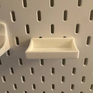 May include: A small, off-white shelf designed for a pegboard system. The shelf is rectangular with a raised lip on all sides, and is mounted on a white pegboard with evenly spaced oval holes. The shelf is empty.