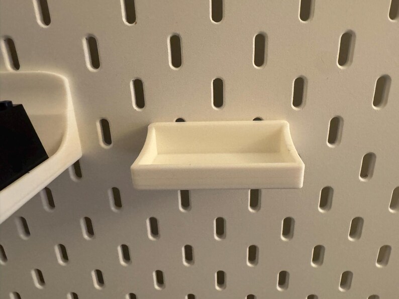 May include: A small, off-white shelf designed for a pegboard system. The shelf is rectangular with a raised lip on all sides, and is mounted on a white pegboard with evenly spaced oval holes. The shelf is empty.