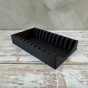 Op de afbeelding: Een zwarte rechthoekige organizer met verticale verdelers. De organizer is gemaakt van een solide materiaal en heeft een gebogen voorrand. Hij staat op een verweerde, lichtgekleurde houten ondergrond.