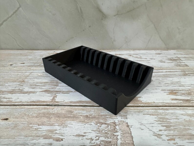 Op de afbeelding: Een zwarte rechthoekige organizer met verticale verdelers. De organizer is gemaakt van een solide materiaal en heeft een gebogen voorrand. Hij staat op een verweerde, lichtgekleurde houten ondergrond.