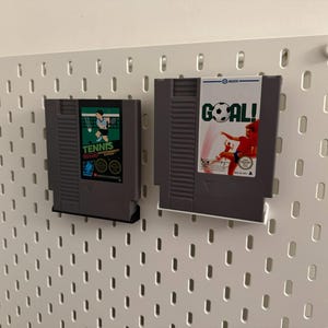 Pode incluir: Dois cartuchos de videojogos retro, um com a etiqueta "Tennis" e o outro "Goal!", s&atilde;o exibidos numa parede perfurada branca. O cartucho "Tennis" apresenta um design verde e preto com um gr&aacute;fico de jogador de t&eacute;nis. O cartucho "Goal!" mostra um jogador de futebol.