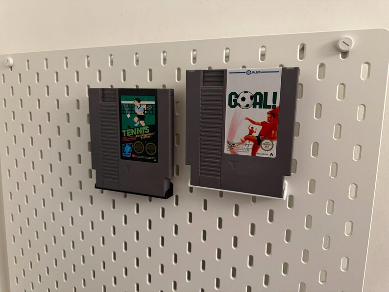 Pode incluir: Dois cartuchos de videojogos retro, um com a etiqueta "Tennis" e o outro "Goal!", s&atilde;o exibidos numa parede perfurada branca. O cartucho "Tennis" apresenta um design verde e preto com um gr&aacute;fico de jogador de t&eacute;nis. O cartucho "Goal!" mostra um jogador de futebol.