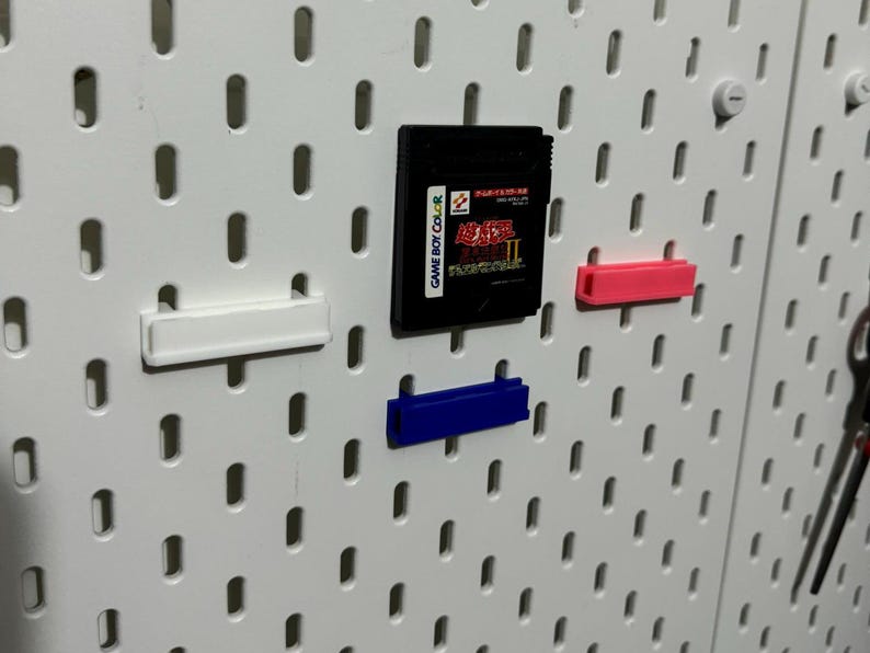 Pode incluir: Um quadro de pinos branco com tr&ecirc;s ganchos de pl&aacute;stico de diferentes cores. Um cartucho de jogo Game Boy Color preto est&aacute; preso ao quadro de pinos. O cartucho de jogo tem o texto "GAME BOY COLOR" e "STREET FIGHTER II" impresso nele.